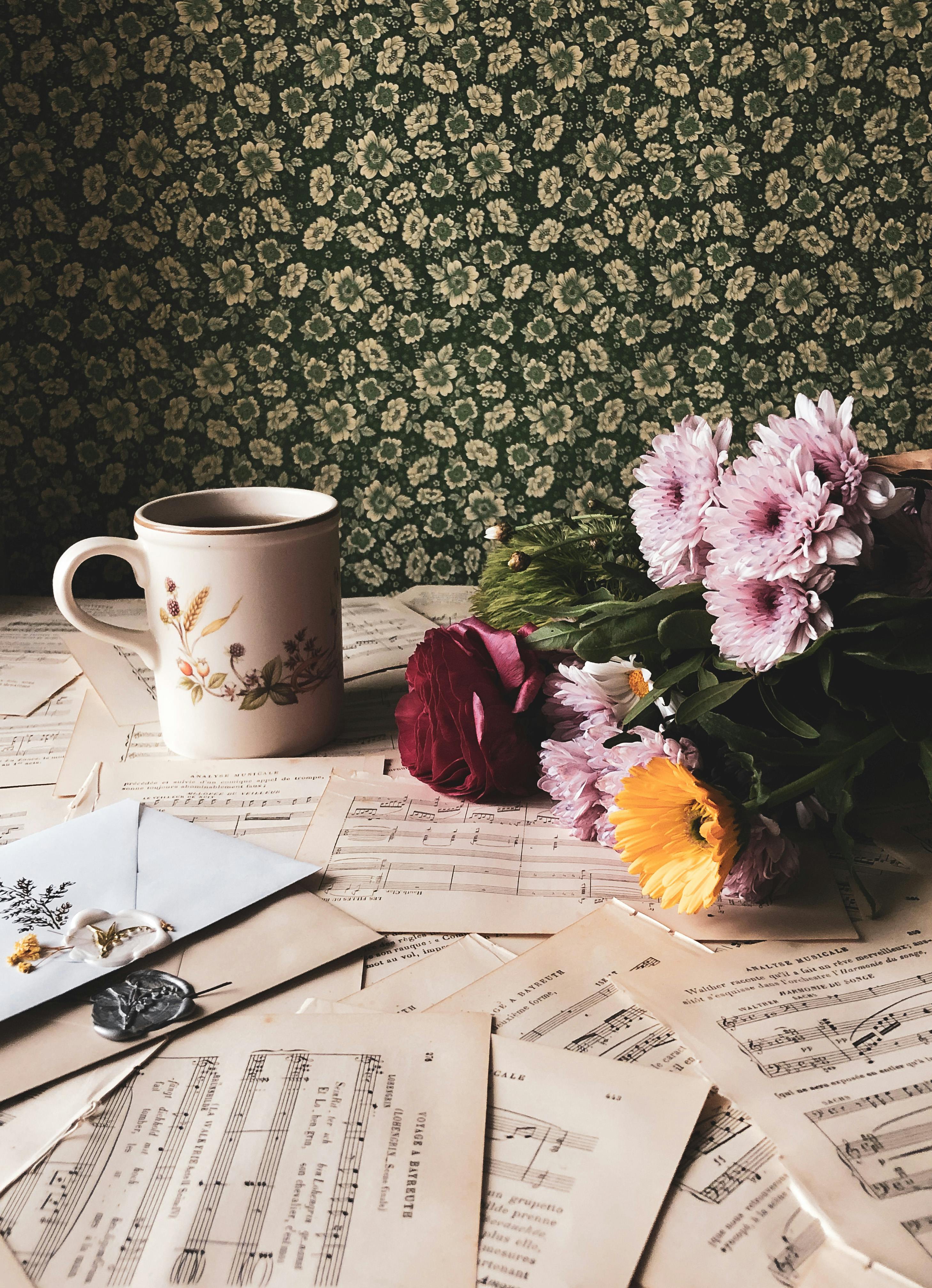 Cup with floral design, bouquet of flowers, and sheet music on a table against a floral-patterned wall.